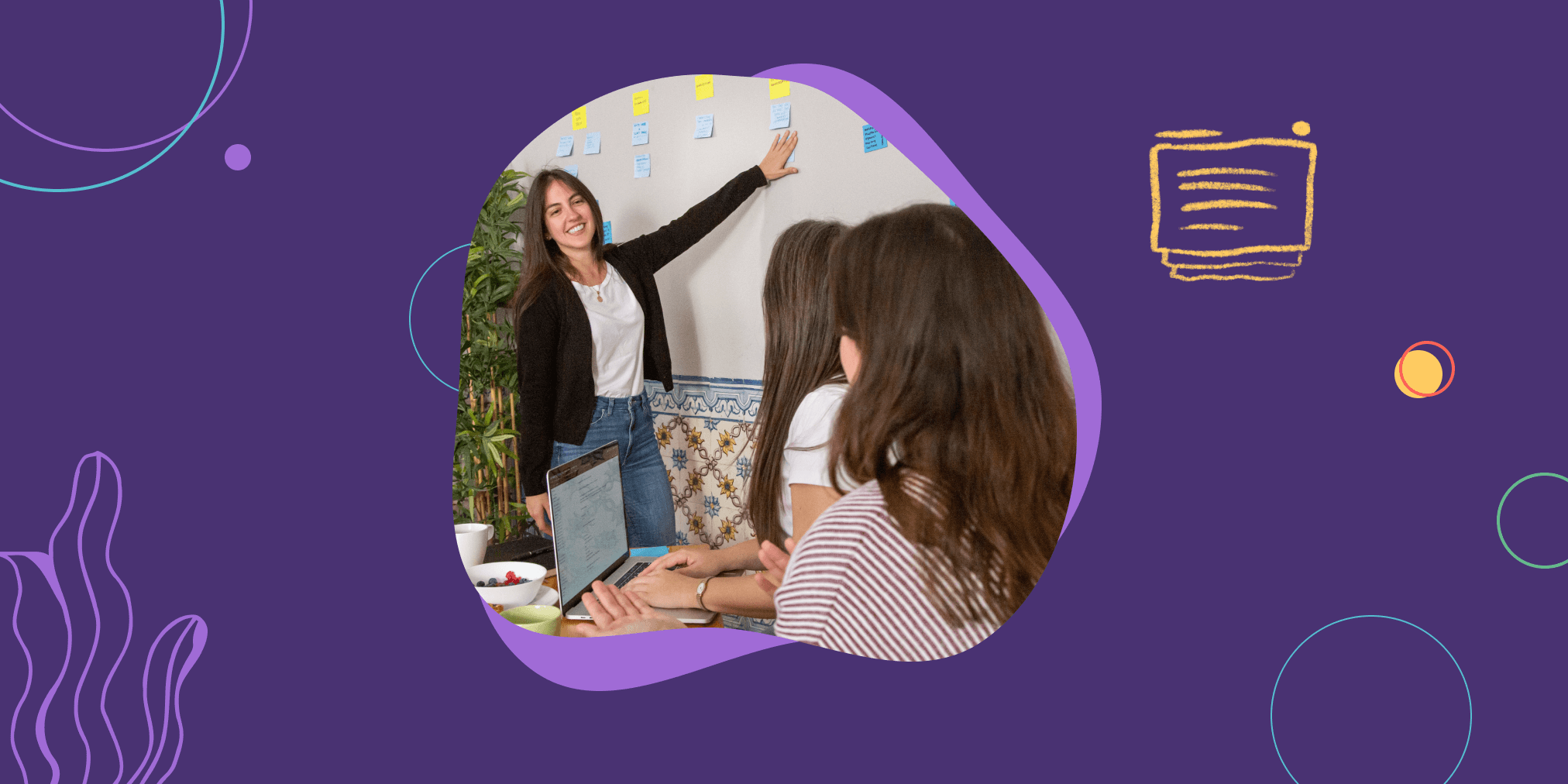 A woman gestures towards a wall with task cards on it in the form of sticky notes, while two team members look on from afar