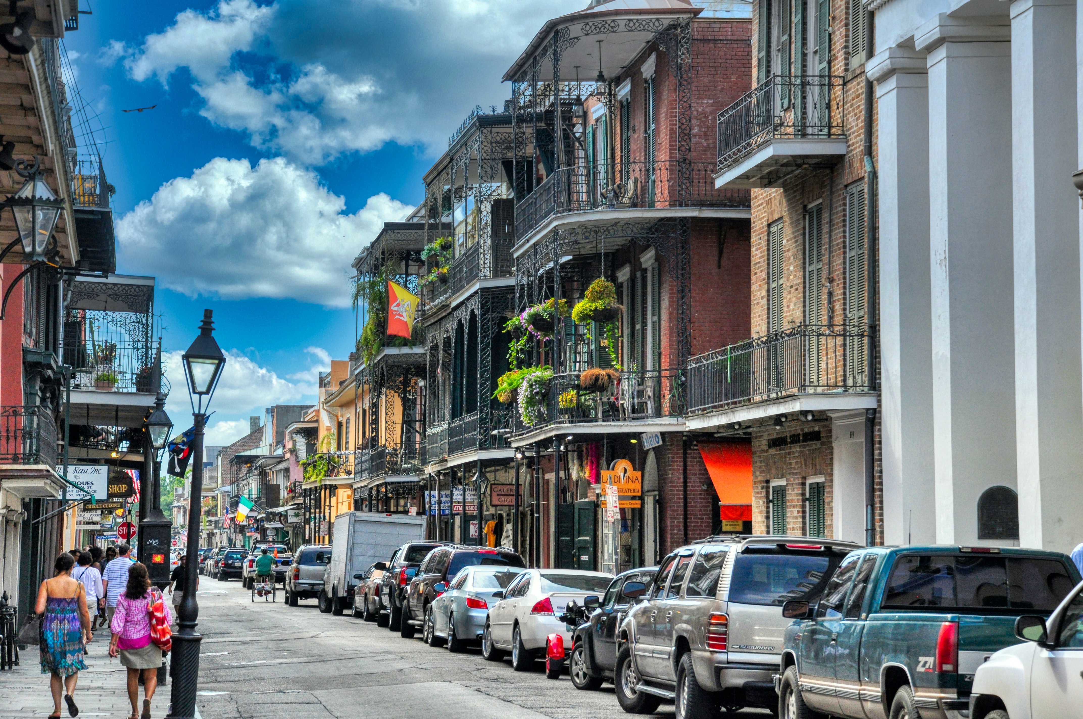 A street from New Orleans’ French Quarter. Colonial-style houses with balconies line the street. Cars are parked on the right side of the street while some people walk on the left side.