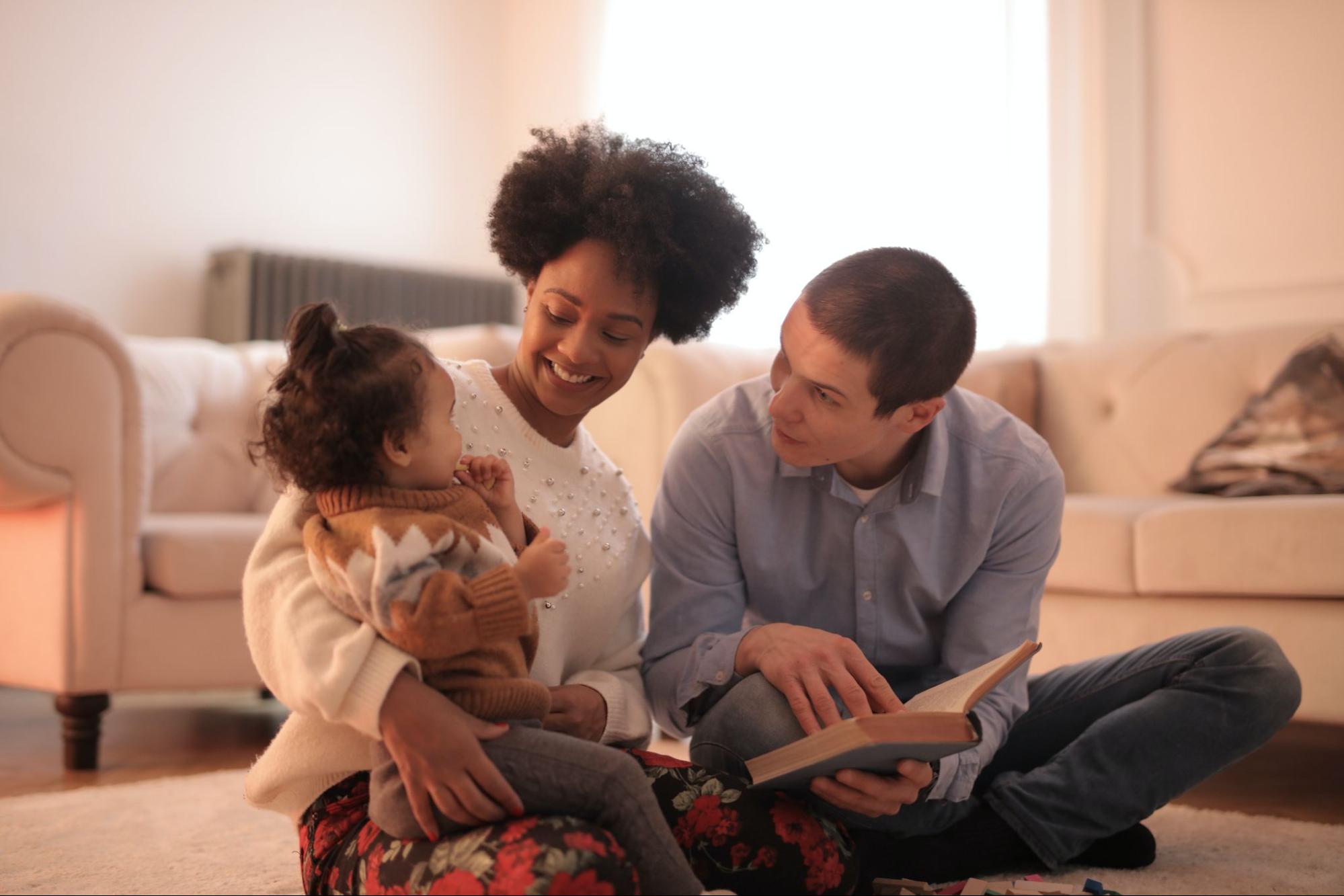 A family sitting on the floor of their living room. A black woman and a white man read a book to their interracial daughter.