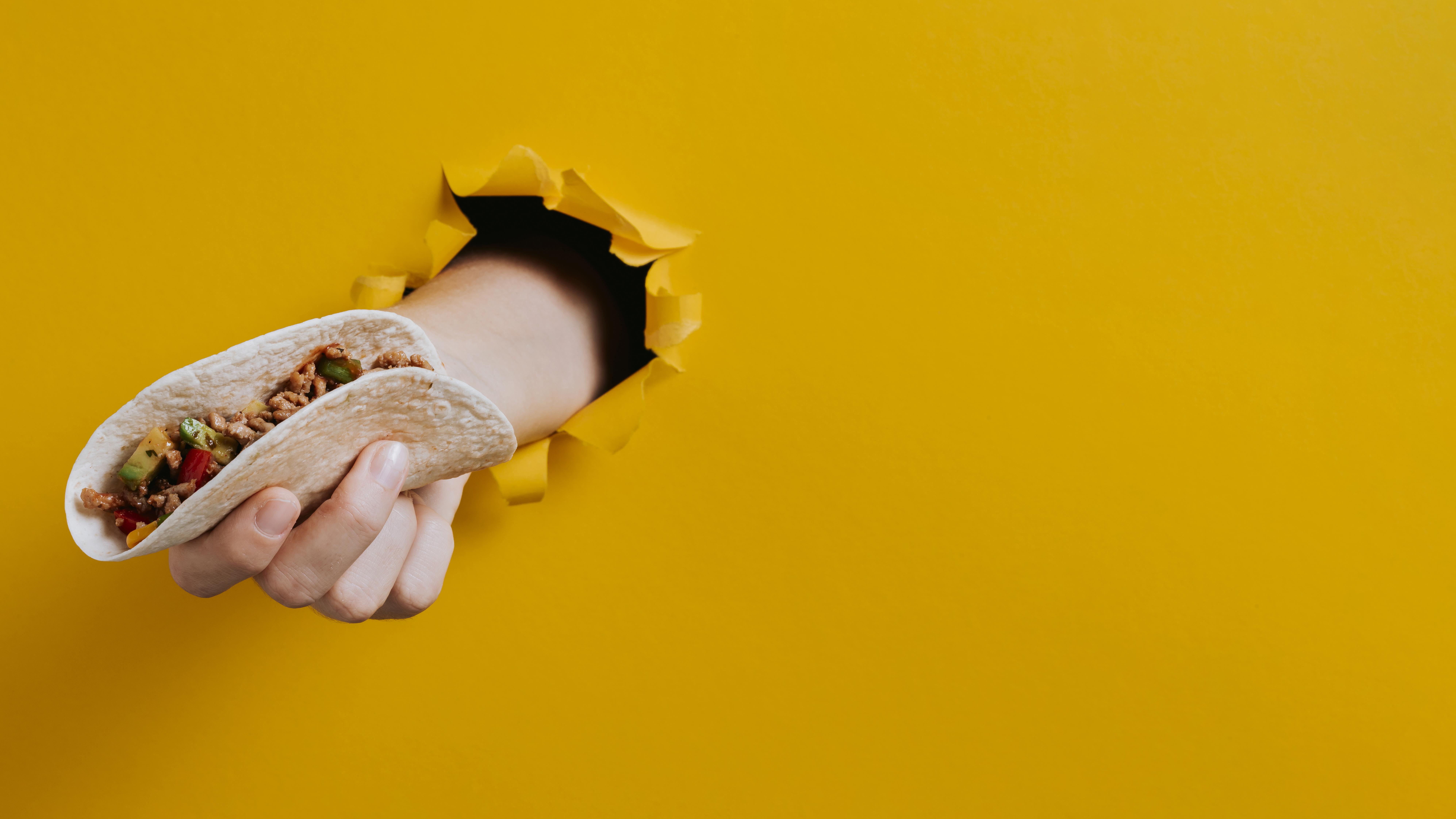 A hand coming out of a yellow wall, holding a taco