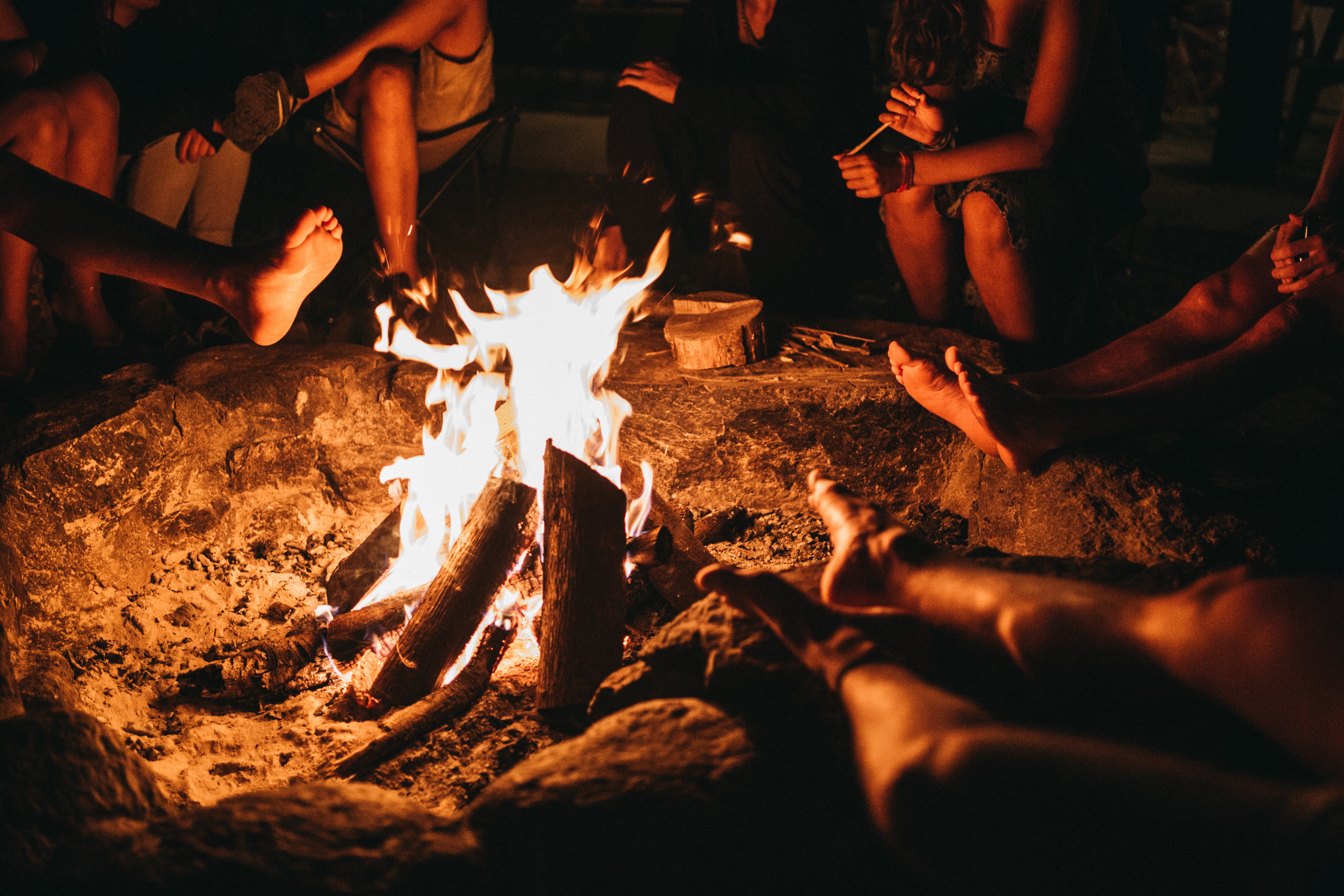 Close up of a group of friends gathered around a bonfire. Only their legs and feet can be seen, and the fire is at the center of the image.